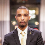 Roland looks toward the camera head on. He's wearing a dark gray suit over a white shirt with a soft yellow tie. The background is a blue of lights.