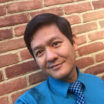 Vu Le gives a close-lipped smile in a selfie in front of a brick wall. He's wearing a bright blue shirt and checkered blue tie.