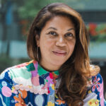 Angelique smiles in her headshot. She's wearing a colorful and whimsical top. She's sitting outside, with the background a blur.